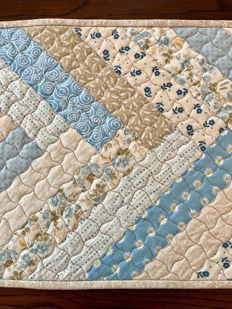 May include: A quilted placemat with a diagonal pattern of various fabrics. The fabrics include light blue with white daisy patterns, cream with small blue floral prints, and tan with floral patterns. The edges are finished with a patterned border.