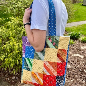 May include: A patchwork tote bag with a variety of colourful fabric squares. The bag has blue straps with white polka dots. The bag is carried over the shoulder.
