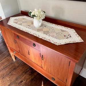 Peut inclure: Un chemin de table matelassé présentant un motif en patchwork composé de divers imprimés floraux et géométriques dans des tons crème, beige et brun doux. Le chemin de table a des extrémités pointues et porte un petit pichet blanc de fleurs.