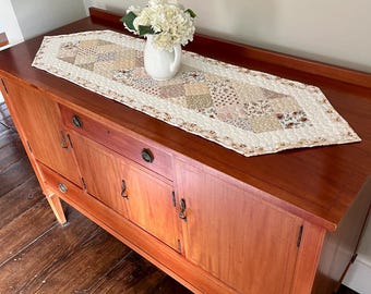 May include: A quilted table runner featuring a patchwork design of various floral and geometric patterns in shades of cream, beige, and muted brown. The runner has pointed ends and is decorated with a small white pitcher filled with white flowers.