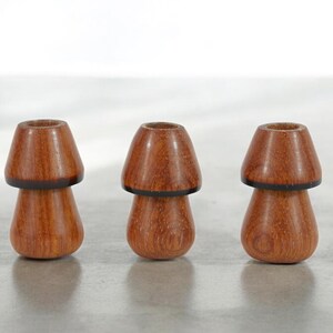 May include: Three wooden mushroom-shaped candle holders. The candle holders are made of dark brown wood with a black band around the top of each.