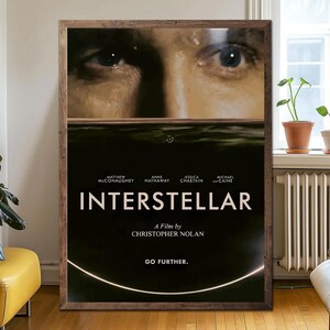 May include: A framed movie poster for "Interstellar" featuring a close-up of a person's eyes above a dark, watery scene. The title is in large white letters, with the names of the actors and director below. The tagline "Go Further" is at the bottom.