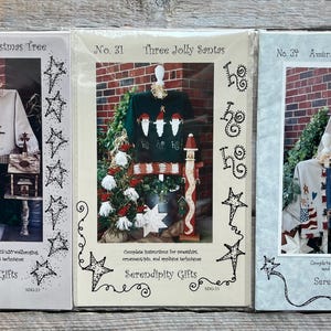 May include: Three vintage craft pattern books featuring holiday sweater designs. The covers display images of finished sweaters with Christmas and patriotic themes. Titles include "Country Christmas Tree," "Three Jolly Santas," and "America! Land That I Love."