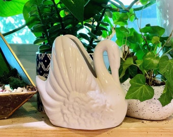 May include: White ceramic swan planter with a decorative design. The planter is sitting on a wooden surface with green plants in the background.