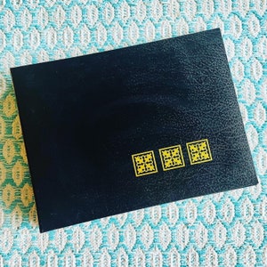 May include: A black leather-bound journal with gold decorative accents. The journal has three gold decorative squares on the front cover.