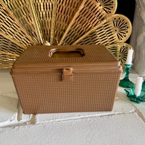 May include: A rectangular, metallic gold storage box with a textured, woven-look exterior. The box features a hinged lid with a recessed handle and a central latch closure. The design is uniform in color and texture across the front and top panels.