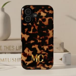 May include: A smartphone case featuring a brown and black tortoiseshell pattern with the gold-colored initials MC printed in the lower center. The case has a glossy finish and a cutout for the camera lens located in the upper left corner.