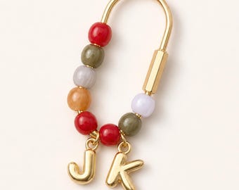 May include: A gold-toned U-shaped earring featuring a series of colorful beads in red, olive green, light gray, and orange. Two gold-toned letter charms, J and K, hang from the bottom of the earring, which is secured by a smooth gold-toned bar.