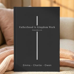 May include: A black hardcover book featuring a white vertical cross graphic. The text reads Fatherhood is Kingdom Work, Proverbs 22:6, and the names Emma, Charlie, and Owen are printed in white at the bottom of the front cover.