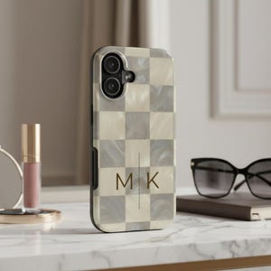 May include: A phone case featuring a checkered pattern of cream and gray marbled squares. The center of the case displays the gold-colored letters M and K separated by a thin vertical line. The case has a black border and a camera cutout.