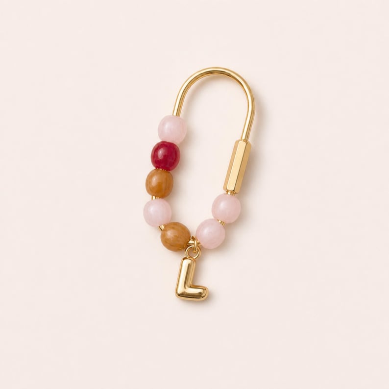 May include: A gold-toned U-shaped earring featuring a series of smooth, round beads in shades of light pink, dark red, and tan. A small, polished gold-toned letter L charm hangs from the bottom of the earring, attached to the metal frame.