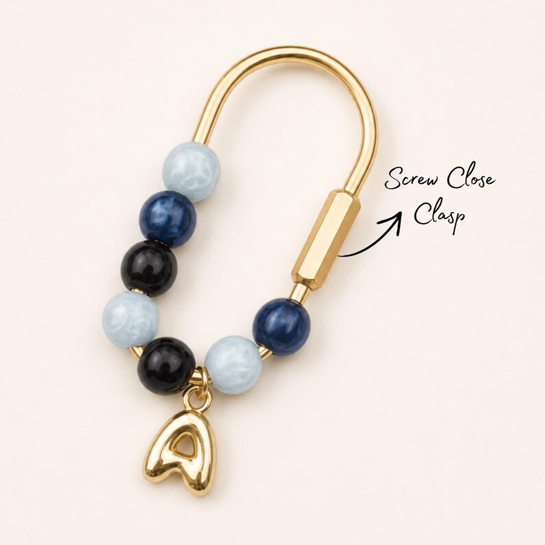May include: A gold-toned U-shaped keychain featuring a screw close clasp and a series of alternating light blue, dark blue, and black beads. A gold-toned letter A charm hangs from the bottom of the keychain. The text Screw Close Clasp is visible.