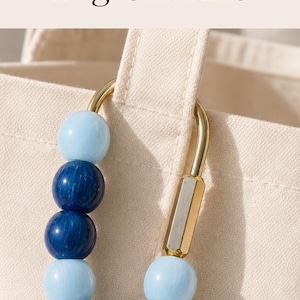 May include: A gold-toned metal carabiner bag charm featuring a variety of colorful beads and a hanging gold letter charm. The accessory is attached to a cream-colored canvas tote bag, which displays the embroidered red letters PBJ on its front.