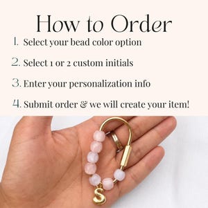 May include: A gold-toned metal carabiner keychain featuring five light pink beads and a single gold-toned letter S charm. The image includes text instructions: How to Order, 1. Select your bead color option, 2. Select 1 or 2 custom initials, 3. Enter your personalization info, 4. Submit order & we will create your item!