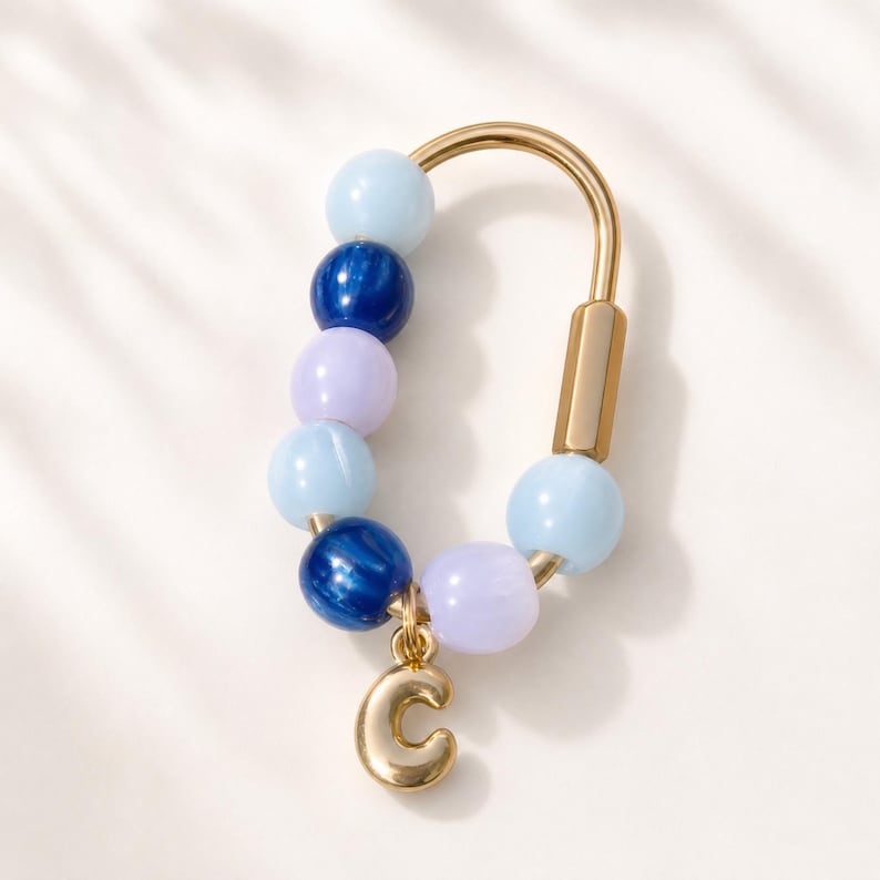 May include: A gold-toned metal carabiner keychain featuring a series of smooth, round beads in shades of light blue, dark blue, and lavender. A small, polished gold-toned letter C charm hangs from the bottom of the carabiner loop.