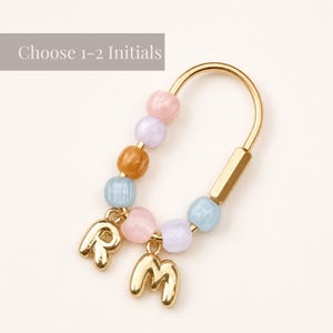 May include: A gold-toned metal keychain featuring a U-shaped frame adorned with pastel-colored beads in pink, lavender, light blue, and tan. Two gold-toned letter charms, R and M, hang from the bottom. The text Choose 1-2 Initials is visible.