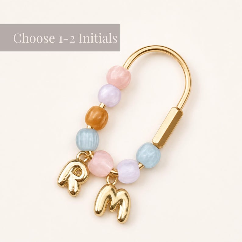 May include: A gold-toned metal keychain featuring a U-shaped frame adorned with pastel-colored beads in pink, lavender, light blue, and tan. Two gold-toned letter charms, R and M, hang from the bottom. The text Choose 1-2 Initials is visible.