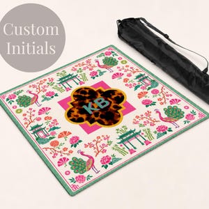 May include: A square mat featuring a white background with a colorful pattern of peacocks, pink flowers, and green pagodas. In the center, a pink square holds a brown leopard print shape with the teal initials KB. A black carrying bag is included.
