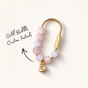May include: A gold-toned metal carabiner keychain featuring a series of light pink and lavender translucent bubble beads threaded along one side, with a gold-toned letter S charm hanging from the bottom. Text reads Gold Bubble Custom Initials.