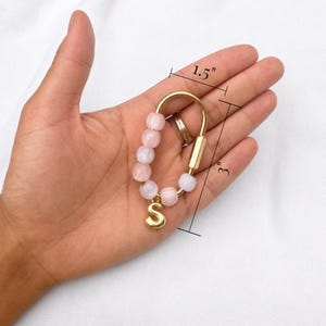 May include: A gold-toned metal carabiner keychain featuring six light pink beads and a gold-toned letter S charm. The keychain measures 1.5 inches in width and 3 inches in length as indicated by the visible measurement lines on the image.