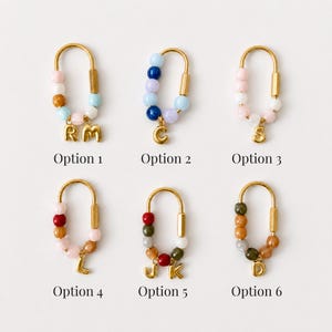 May include: Six gold-toned U-shaped carabiner charms decorated with various colored beads and gold letter pendants. Option 1 features letters R and M, Option 2 has letter C, Option 3 has letter S, Option 4 has letter L, Option 5 has letters J and K, and Option 6 has letter D.
