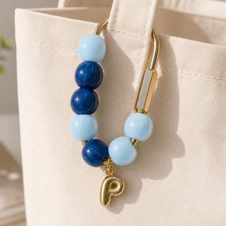 May include: A gold-toned metal carabiner keychain featuring a series of alternating light blue and dark blue spherical beads, with a gold-toned letter P charm hanging from the bottom of the loop, attached to a cream-colored canvas bag.