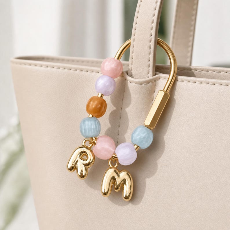 May include: A gold-toned metal carabiner-style bag charm featuring a series of smooth, round beads in pastel pink, lavender, amber, and light blue. Two polished gold-toned metal letter charms, R and M, hang from the bottom of the bead strand.