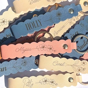 May include: A collection of scalloped-edge keychains in pastel blue, peach, and cream colors. Each keychain features a metal snap, a silver-toned ring, and is engraved with a unique name and a delicate floral line art illustration.