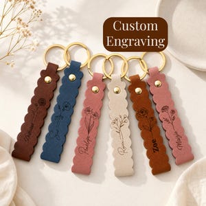 May include: Six scalloped leather keychains in dark brown, blue, pink, cream, tan, and dusty rose. Each features a gold-tone metal ring, a gold rivet, and a black engraved floral design with a name. The text Custom Engraving is visible.