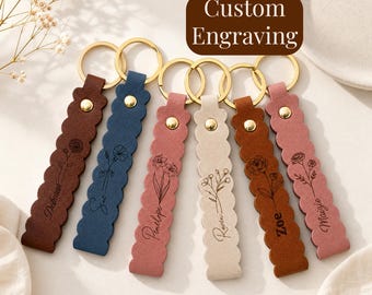 May include: Six scalloped leather keychains in dark brown, blue, dusty pink, cream, tan, and mauve, each featuring a gold-tone metal ring, a gold rivet, and a custom engraving of a floral illustration with a name. The text Custom Engraving is visible.