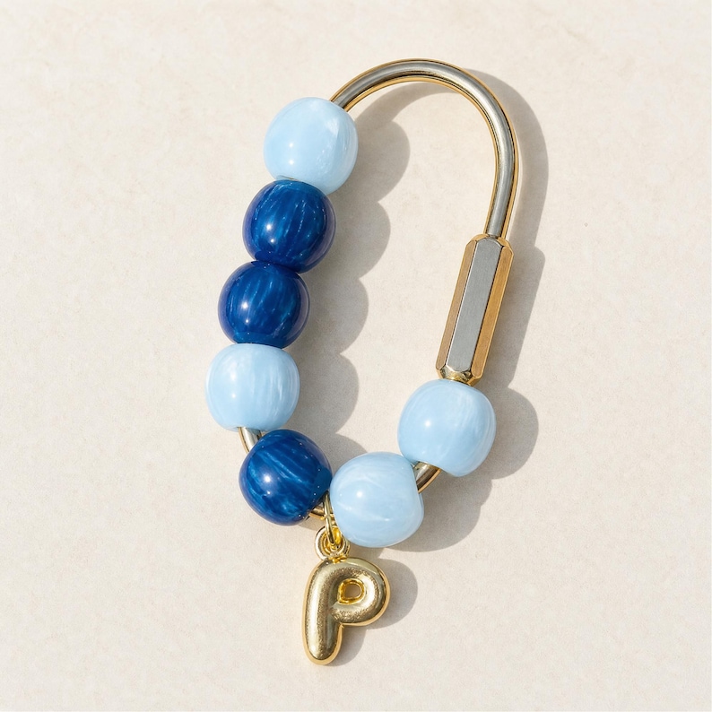 May include: A gold-toned metal carabiner keychain featuring a series of alternating light blue and dark blue spherical beads. A small, gold-toned metal charm in the shape of the uppercase letter P hangs from the bottom of the carabiner frame.