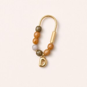May include: A gold-toned U-shaped carabiner keychain featuring a series of smooth, round beads in shades of olive green, amber, and light gray. A polished gold-toned letter D charm hangs from the bottom of the carabiner, secured by a small ring.