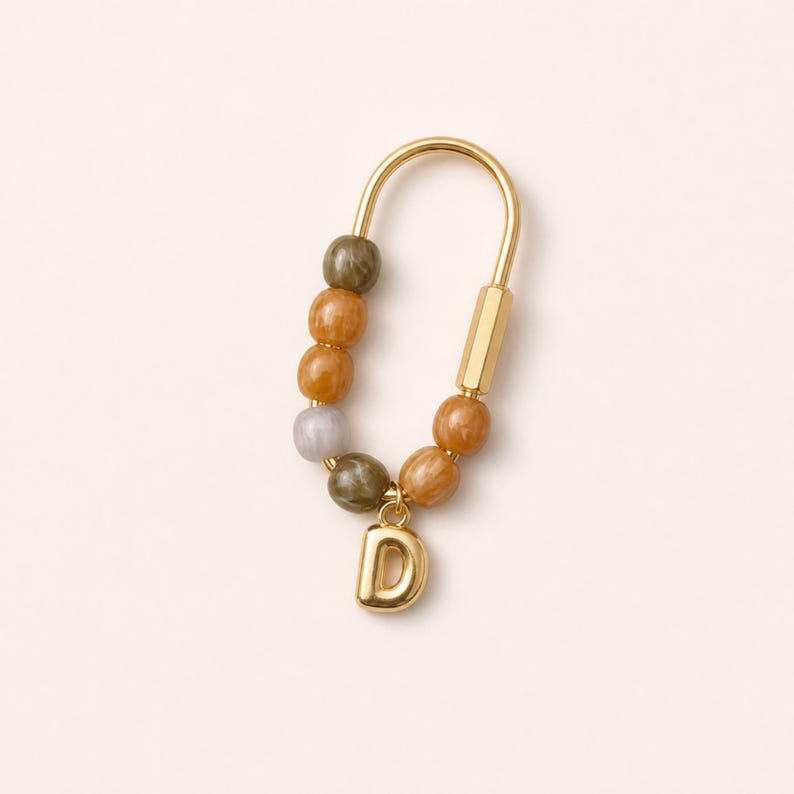 May include: A gold-toned U-shaped carabiner keychain featuring a series of smooth, round beads in shades of olive green, amber, and light gray. A polished gold-toned letter D charm hangs from the bottom of the carabiner, secured by a small ring.