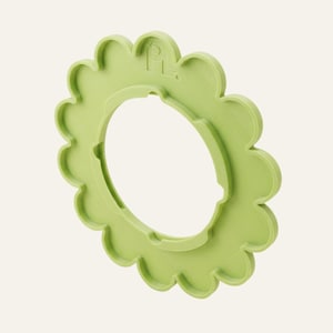 May include: A light green, flower-shaped plastic attachment with a scalloped edge and a circular centre opening. The object features a small, embossed logo at the top that reads PL with a small circle symbol, showing a flat, matte finish.