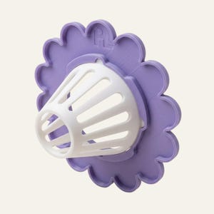 May include: A lavender flower-shaped base with a scalloped edge and a white, conical, slotted basket attachment protruding from the centre. The base features a small embossed logo at the top, and the entire object has a matte finish.