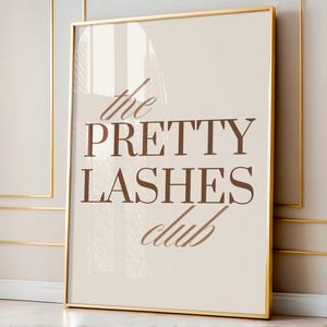 May include: A framed print featuring the words "the PRETTY LASHES club" in brown script on a cream backdrop. The frame is gold, and the print is displayed against a neutral wall.