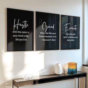 Puede incluir: Tres impresiones artísticas de pared negras enmarcadas con escritura blanca. Las impresiones dicen "Hustle", "Grind" y "Execute" con texto motivacional. Una consola negra sostiene un jarrón blanco, una roca y un vaso decorativo con tonos azules y amarillos.
