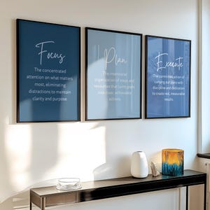 Puede incluir: Tres carteles motivacionales enmarcados con las palabras "Focus", "Plan" y "Execute". Cada cartel tiene un fondo azul diferente. Una consola negra sostiene un jarrón, una piedra decorativa y un portavelas de cristal.