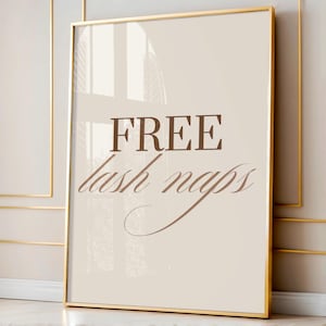 Puede incluir: Un estampado enmarcado con un borde dorado. El estampado tiene un fondo color crema con las palabras "FREE lash naps" en letras marrones y en bloque. El estampado se muestra contra una pared neutra.