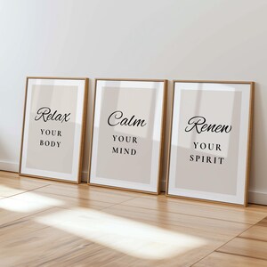 May include: Three framed prints with the words "Relax Your Body", "Calm Your Mind", and "Renew Your Spirit" in black script. The prints have a beige background and are in wooden frames, leaning against a white wall. The floor is wooden.