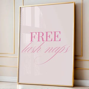 Puede incluir: Un estampado enmarcado con un borde dorado. El estampado tiene un fondo rosa claro con las palabras "FREE" y "lash naps" en escritura rosa. El estampado está apoyado contra una pared.