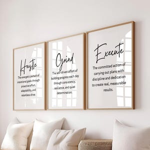 May include: Three vertical wall art prints in light wood frames featuring white backgrounds with black text. The prints display the words Hustle, Grind, and Execute, each followed by a descriptive definition of the term in a clean, sans-serif font.