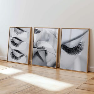 May include: A set of three framed black and white prints. Each print features a close-up of eyelashes. The prints are in wooden frames and are displayed against a white wall. The images capture the detail of the eyelashes.