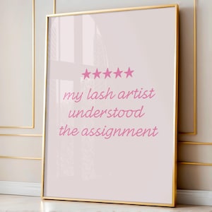 May include: A framed print with a gold border. The print has a light pink background with the text "my lash artist understood the assignment" in pink script. Above the text are five pink stars.
