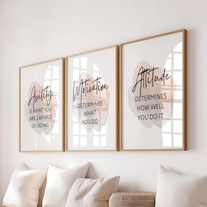 Op de afbeelding: Een set van drie ingelijste wandkunstprints met lichte houten lijsten. Elke print bevat een zachtroze abstracte vorm in aquarel-stijl achter zwarte tekst. De tekst luidt: Ability is what you are capable of doing, Motivation determines what you do en Attitude determines how well you do it.