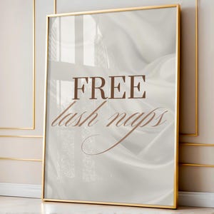 Puede incluir: Un estampado enmarcado con un marco dorado. El estampado presenta las palabras "FREE lash naps" en escritura marrón sobre un fondo suave y fluido, blanquecino. El marco es rectangular y tiene un acabado brillante.