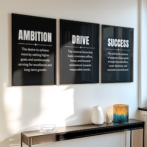 May include: Three framed black and white motivational posters with the words "AMBITION", "DRIVE", and "SUCCESS". Each poster has a definition below the title. A black console table holds a white vase, a small rock, and a decorative glass.