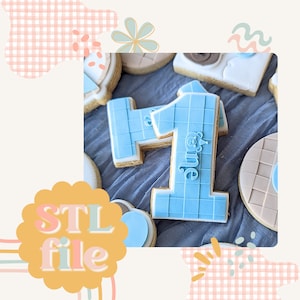 May include: A selection of decorated biscuits, including a large blue number one biscuit with a grid pattern and the word "one". Other biscuits feature various shapes and designs, with a soft colour palette.