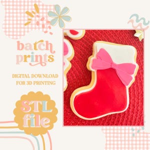 May include: A festive Christmas stocking cookie, featuring a red base, white cuff, and a pink bow. The cookie is outlined in yellow and rests on a red textured surface. The image includes the text "batch prints" and "STL file".