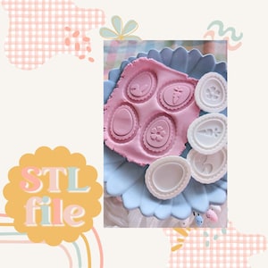 Op de afbeelding: Een set van zes witte plastic paaseierenkoekjesvormen. De vormen zijn gevormd als eieren met verschillende ontwerpen, waaronder een bloem, een konijn en een wortel. De vormen staan op een blauw bord met roze klei op de achtergrond.